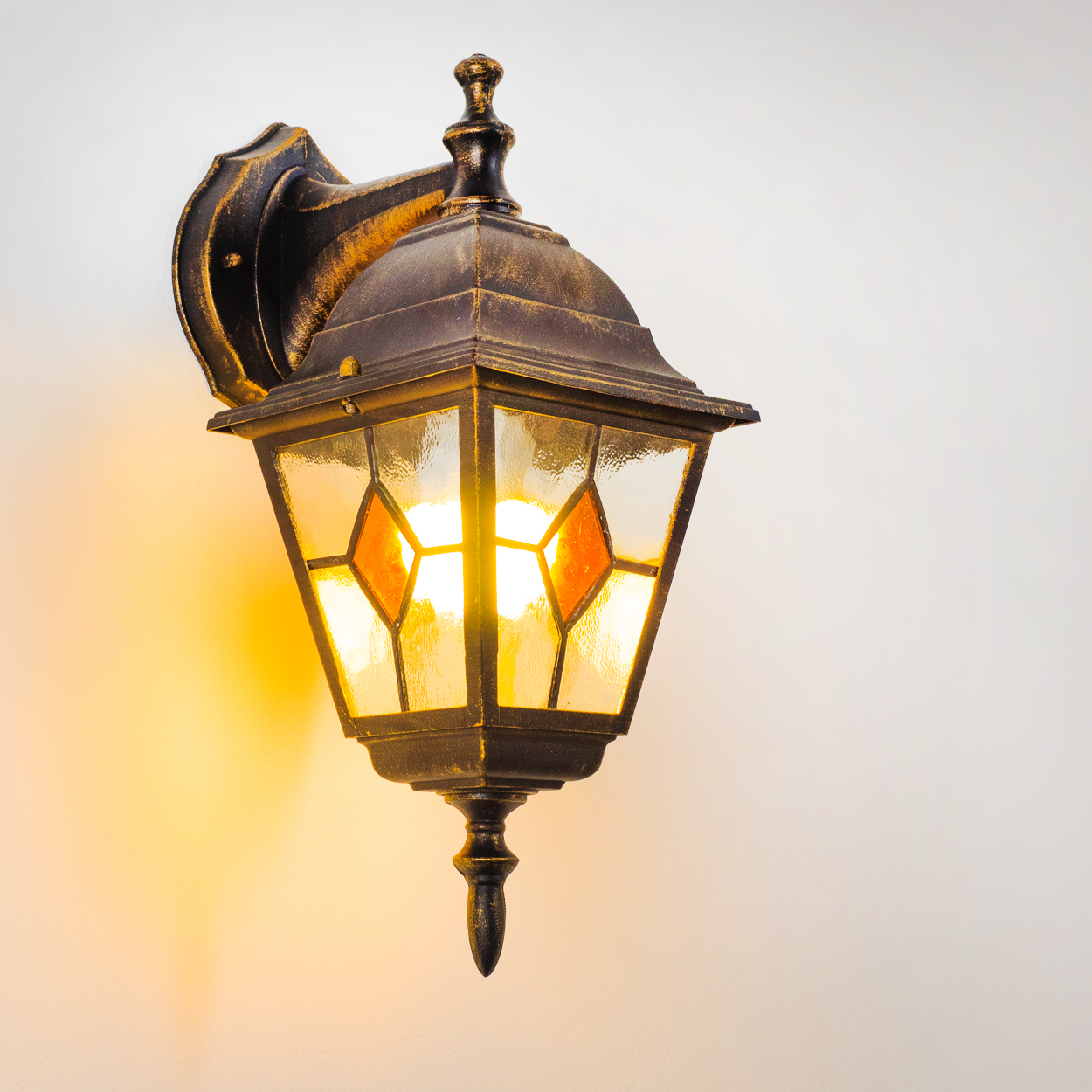 Eine verzierte, beleuchtete Außenlampe mit Glasgehäuse und orangefarbenen Diamantakzenten, an einer Wand befestigt. Der Hintergrund ist weiß.