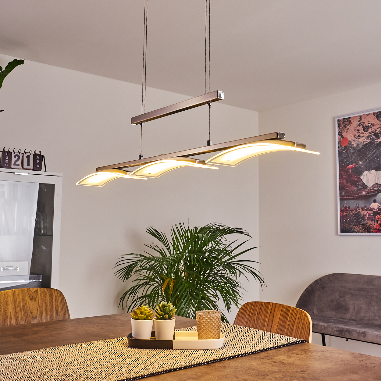 Esszimmer-Szene. Eine moderne Leuchte hängt über einem Tisch mit Pflanzen und einem Läufer. Ein Kalender und Kunst sind sichtbar.