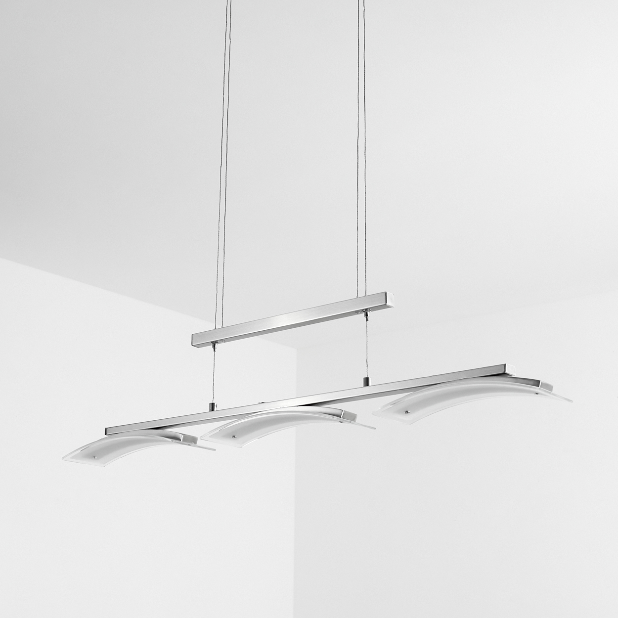 Eine moderne, silberne Deckenleuchte mit drei gebogenen Glasschirmen. Sie hängt an dünnen Drähten vor einer weißen Wand und Decke.