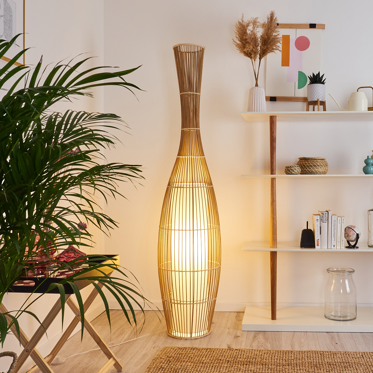 Eine hohe Bambuslampe beleuchtet einen Raum mit einer Zimmerpflanze links und Regalen mit Dekoration rechts.