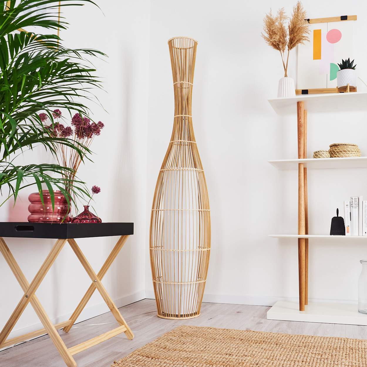 Innenraum mit hoher gewebter Lampe, Pflanzen auf einem Tisch und Regal mit Dekoration vor weißer Wand.