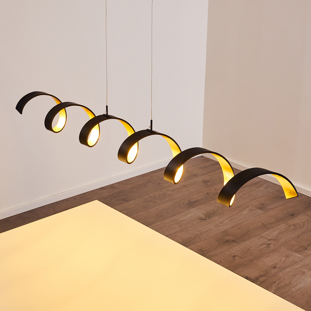 Vier schwarze, spiralförmige Pendelleuchten mit goldenen Innenflächen. Sie hängen über einem Holzboden und einer weißen, beleuchteten Fläche.