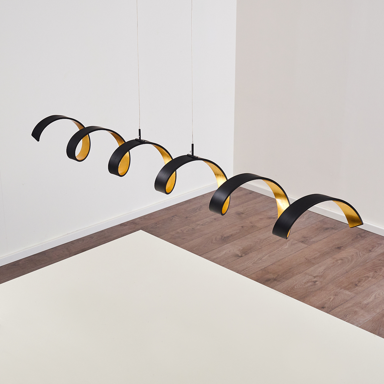 Drei schwarz-goldene Spiral-Pendelleuchten hängen über einem weißen Tisch. Holzboden und weiße Wände vervollständigen die Szene.