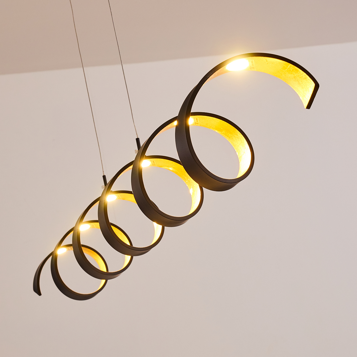 Eine moderne Spiral-Pendelleuchte hängt von der Decke. Sie hat einen schwarzen Rahmen mit goldenem Innenraum und strahlt warmes Licht aus.