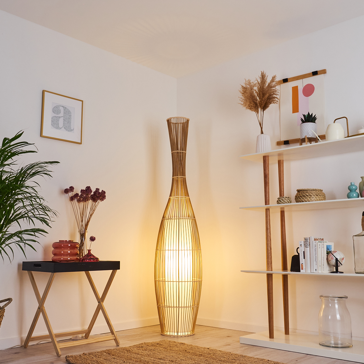 Eine hohe Stehlampe aus Bambus beleuchtet eine gemütliche Ecke. Ein kleiner Tisch mit Blumen und ein Regal mit Dekoration vervollständigen die Szene.
