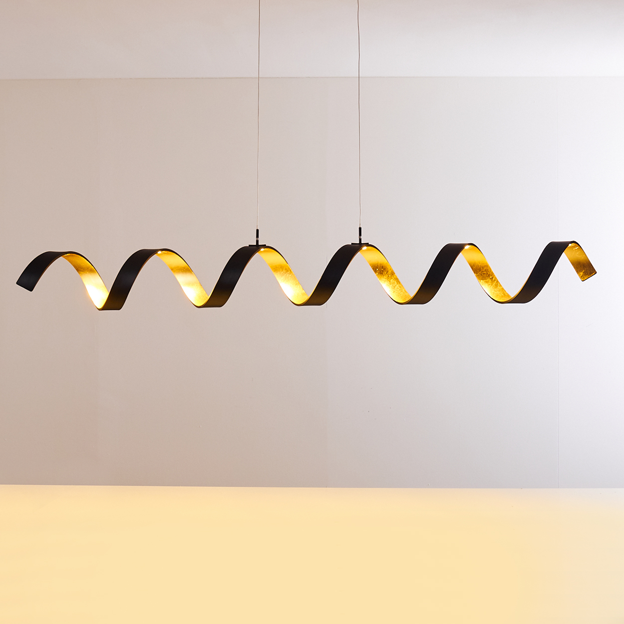 Eine wellenförmige, schwarz-goldene Leuchte hängt von der Decke. Das Design der Leuchte ist eine durchgehende Wellenform.