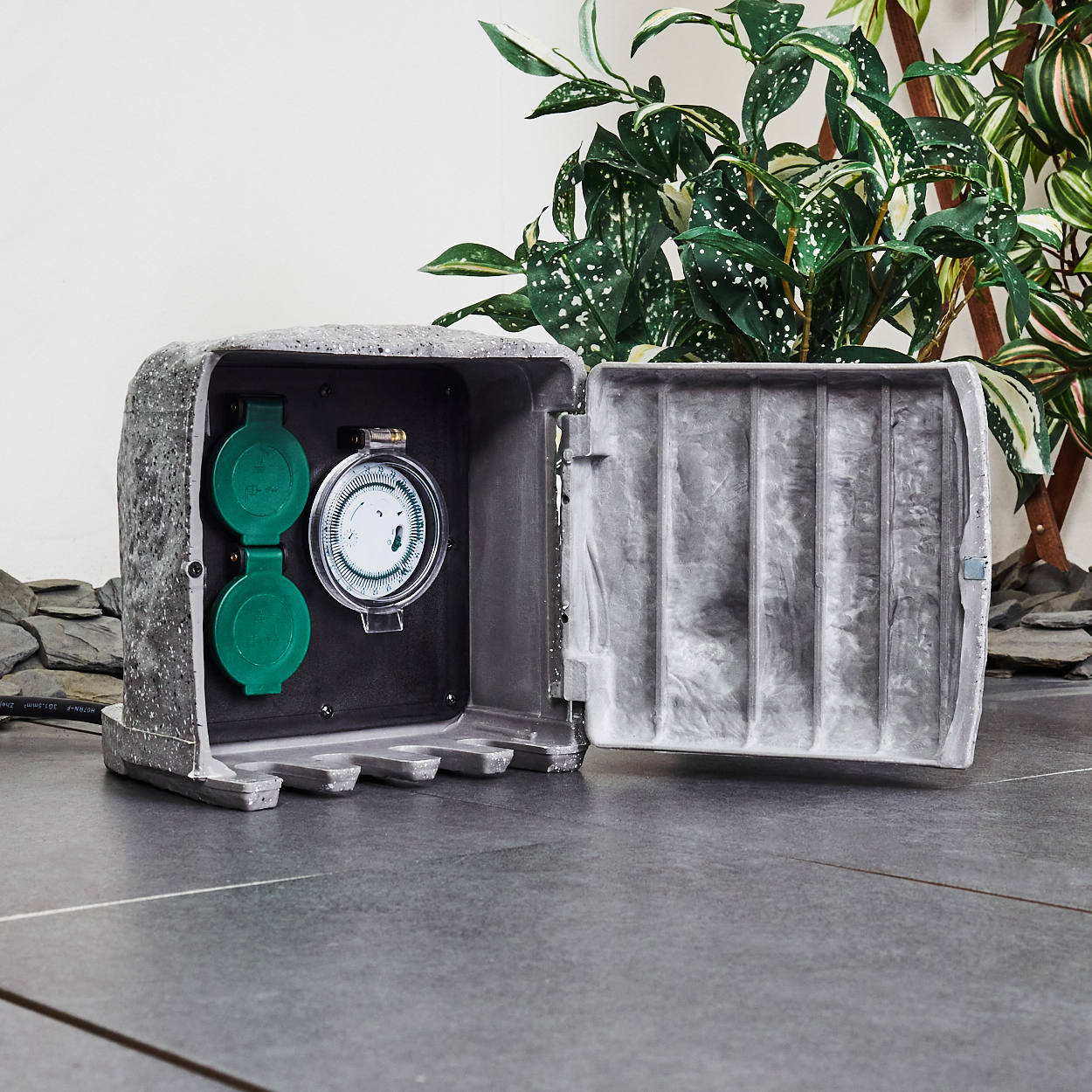 Geöffnete graue Elektrokiste mit grünen Steckdosen und einem Timer, auf dunkler Fliesenoberfläche mit Laub im Hintergrund.
