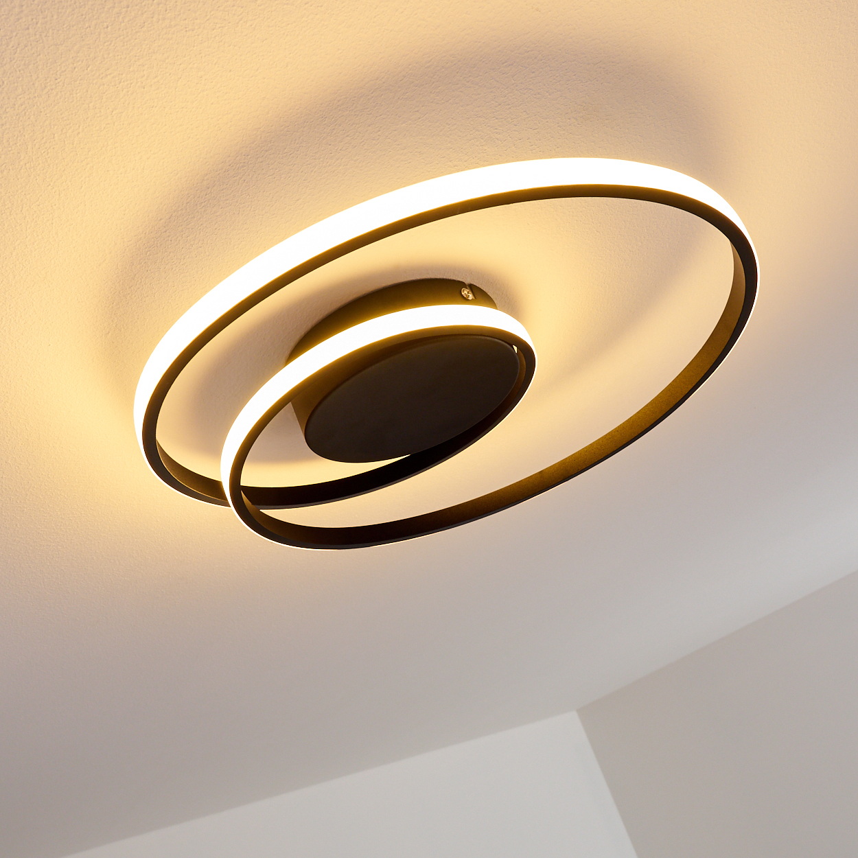 Eine moderne Deckenleuchte mit schwarzem Sockel und zwei leuchtenden, wirbelnden Ringen, die ein warmes Licht auf die weiße Decke werfen.
