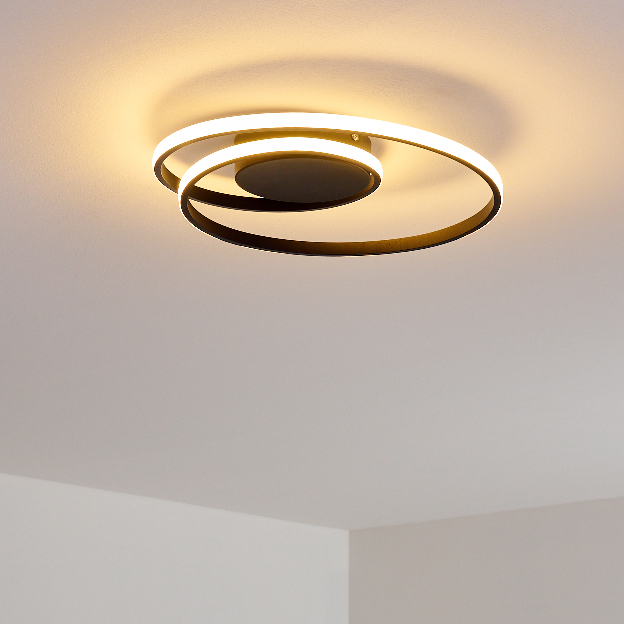 Eine moderne, spiralförmige Deckenleuchte mit schwarzem Rahmen und hellem, weißem Licht. Sie wirft einen Schatten an die weiße Decke.
