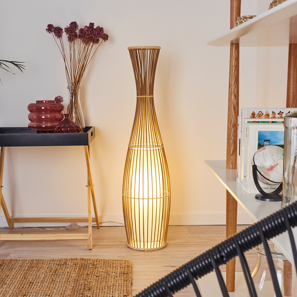 Eine hohe Bambuslampe beleuchtet einen Raum. Ein schwarzes Tablett mit Vasen befindet sich links. Ein Regal mit Büchern befindet sich rechts.