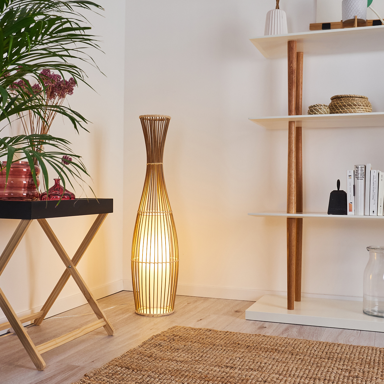 Eine beleuchtete Bambuslampe neben einem Tisch mit Pflanzen und einem Regal mit Büchern. Ein Jute-Teppich auf dem Boden.