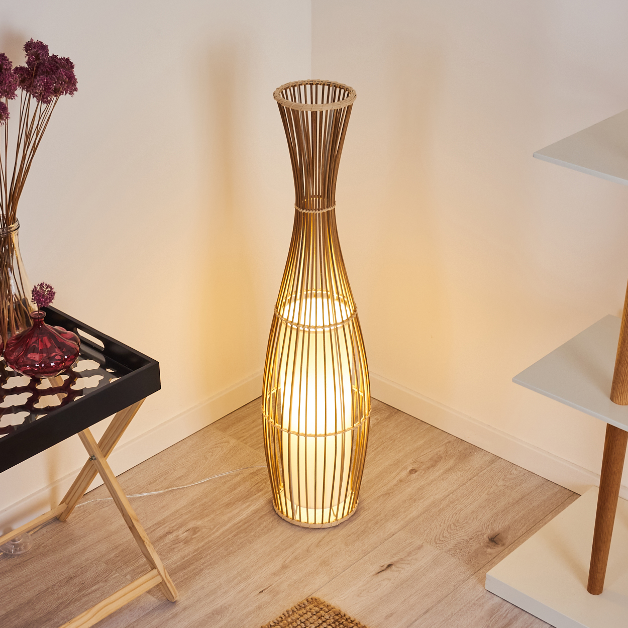 Eine Bambuslampe steht in einer Ecke. Ein schwarzes Tablett mit Blumen und ein weißes Regal sind in der Nähe. Holzboden und weiße Wände.