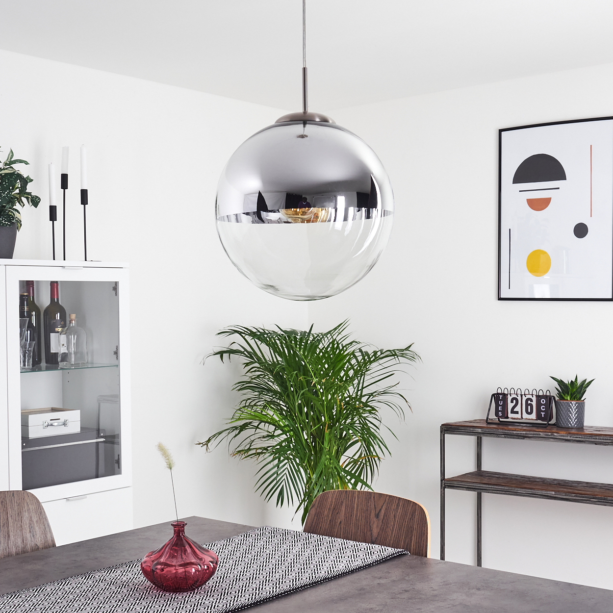 Ein Raum mit Esstisch, Kugel-Lampe und Pflanzen. Ein Schrank mit Weinflaschen und ein gerahmtes abstraktes Kunstwerk.