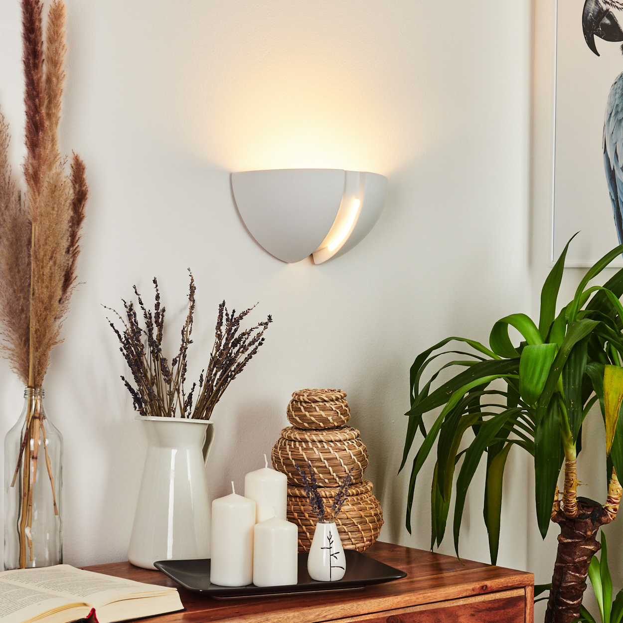 Eine Wandlampe beleuchtet einen Tisch mit Blumen, Kerzen und geflochtenen Körben. Eine Pflanze und ein gerahmtes Vogelkunstwerk sind ebenfalls sichtbar.