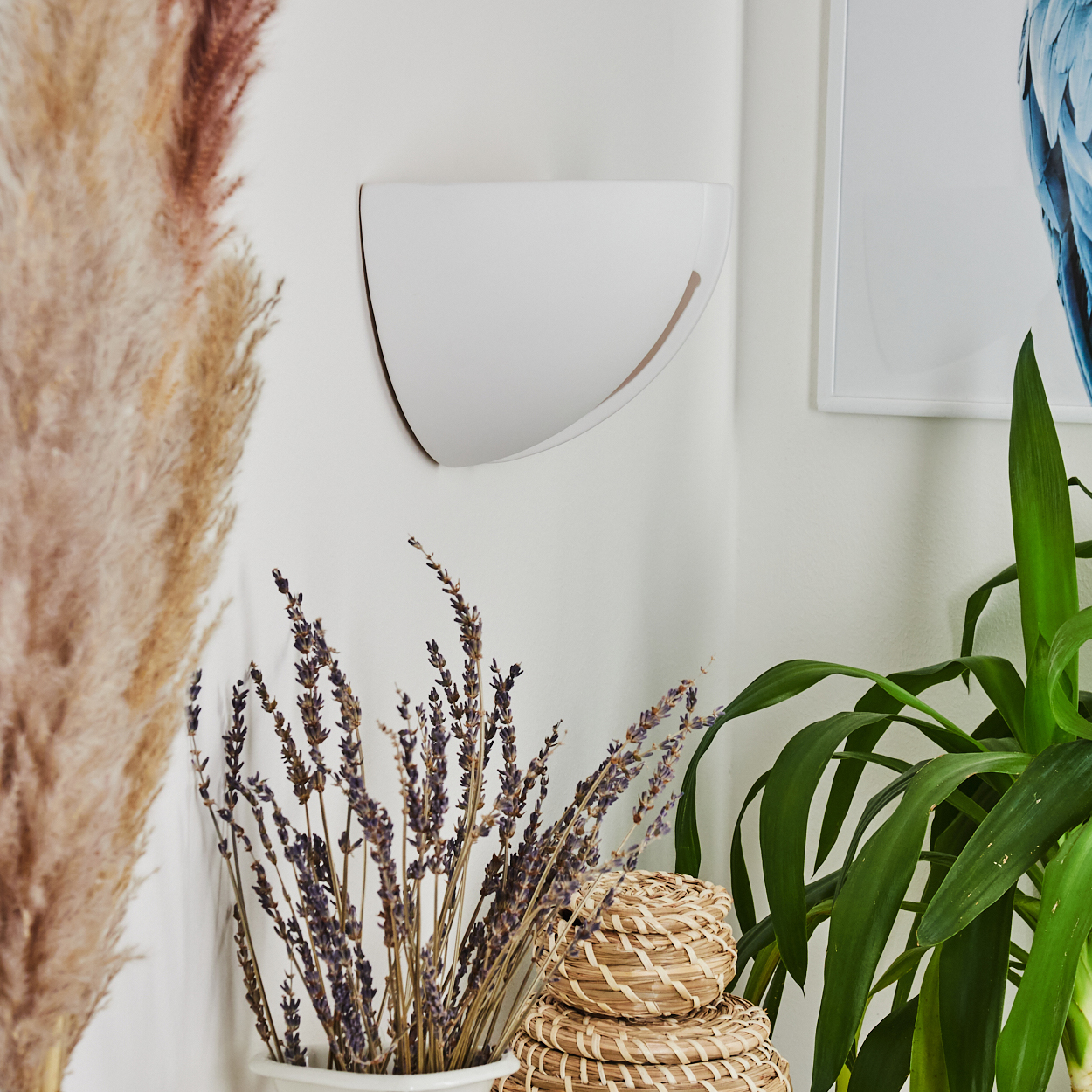 Eine weiße Wandlampe mit goldenem Umriss. Getrockneter Lavendel, Korbflechtkörbe und grüne Pflanzen im Vordergrund. Ein gerahmtes Bild ist sichtbar.