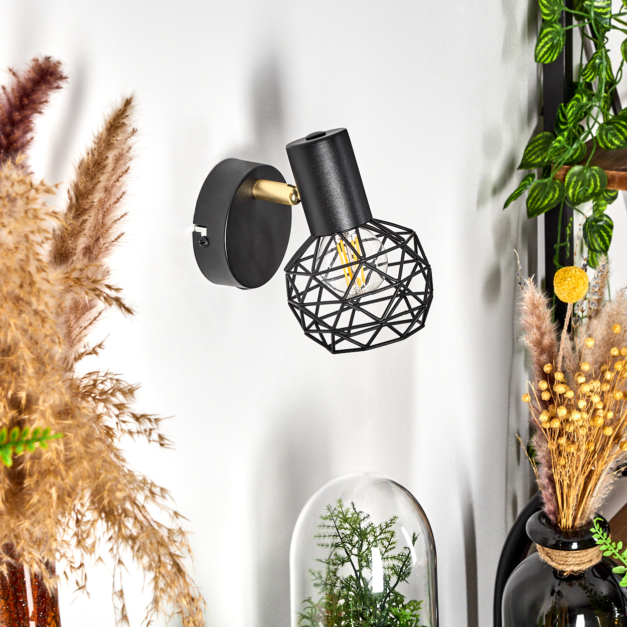 Eine schwarze Wandlampe mit geometrischem Schirm ist an einer weißen Wand befestigt. Trockene Pflanzen und grünes Laub schmücken die Umgebung.