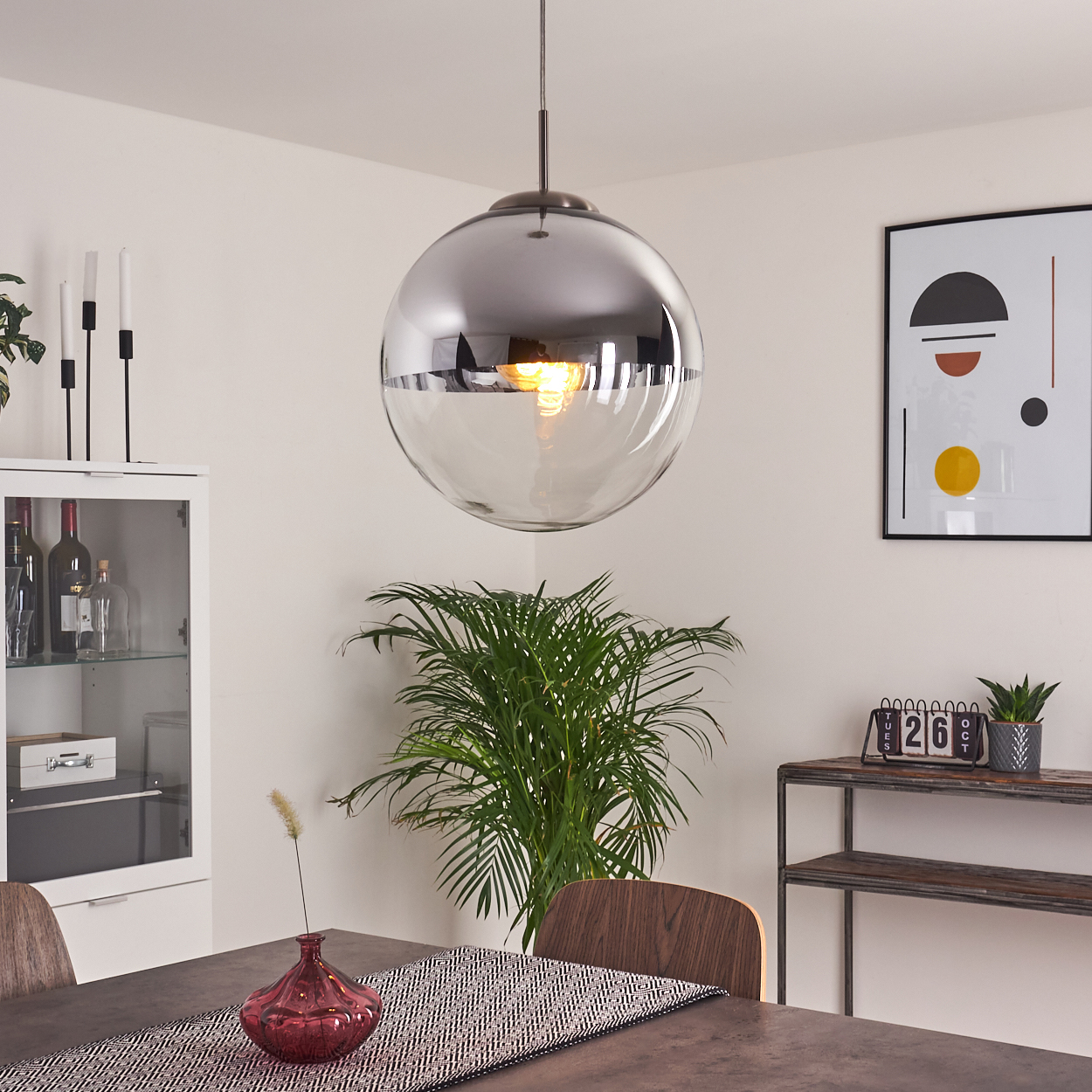 Ein Esszimmer mit einer großen Pendelleuchte, einem Tisch mit Tischläufer und Vase sowie einem gerahmten Kunstdruck an der Wand.