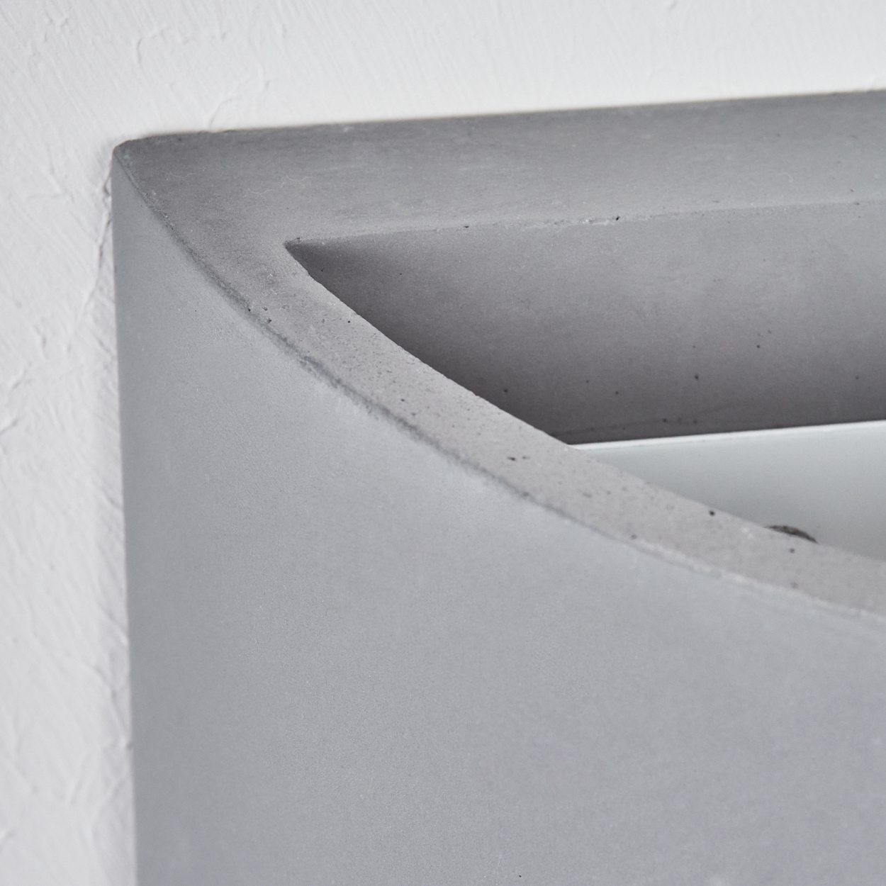 Nahaufnahme eines grauen Betonpflanzgefäßes vor einer weißen Wand. Der Pflanzkasten hat eine gebogene Oberseite und eine rechteckige Öffnung.