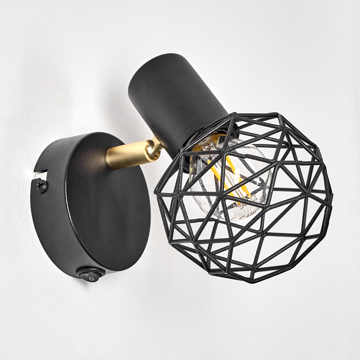 Schwarze Wandleuchte mit geometrischem Schirm, goldenen Akzenten und einer klaren Glühbirne vor weißer Wand. Der Schalter ist sichtbar.