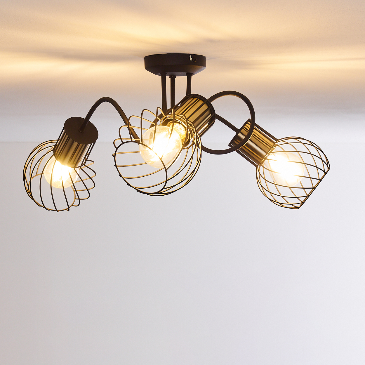 Deckenleuchte mit drei Lampen. Jede Lampe hat einen Drahtkäfig und eine helle Glühbirne. Schwarzer Rahmen an einer weißen Decke.
