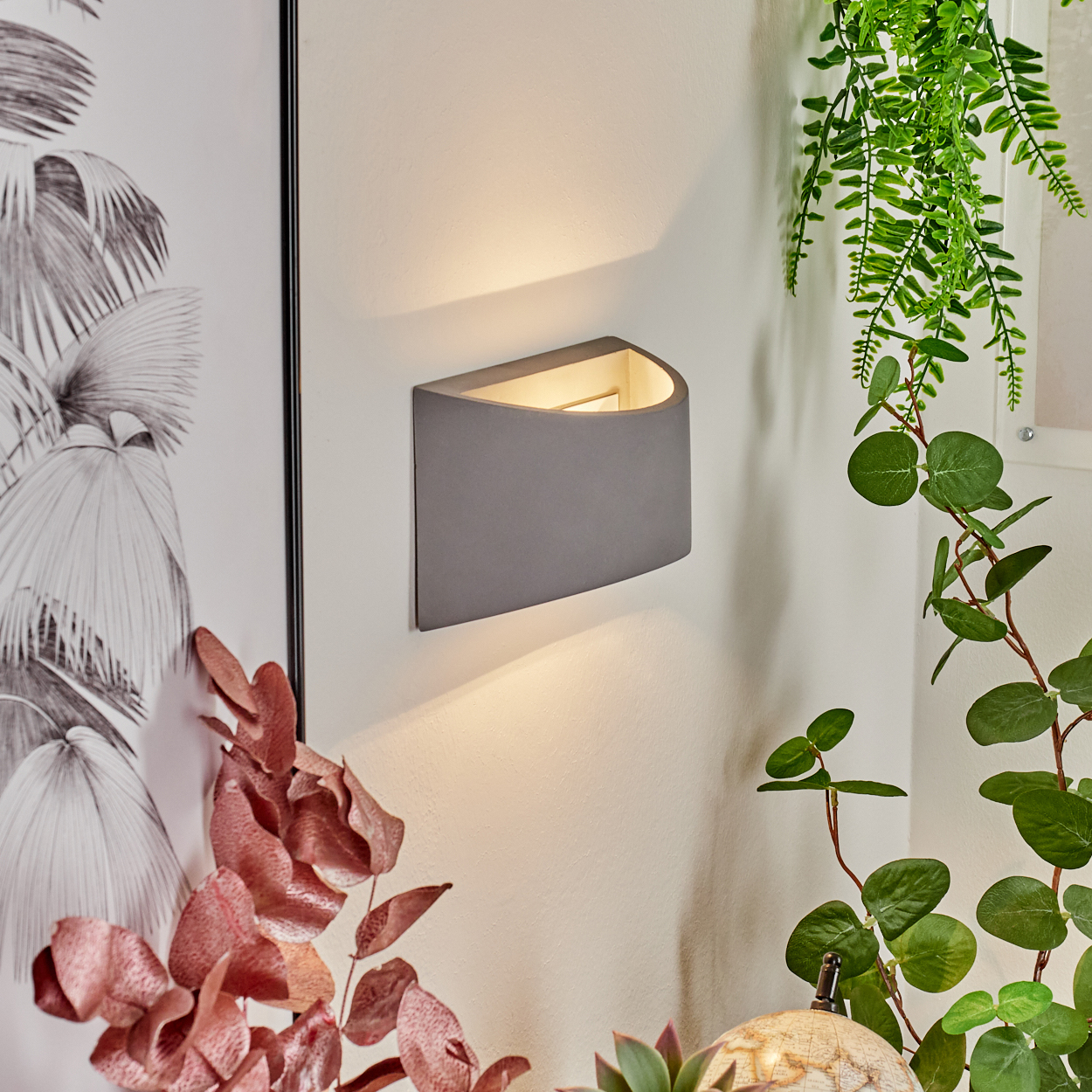 Eine graue Wandleuchte beleuchtet eine weiße Wand. Grüne und rötliche Pflanzen umgeben die Lampe und ein gerahmtes Kunstwerk.