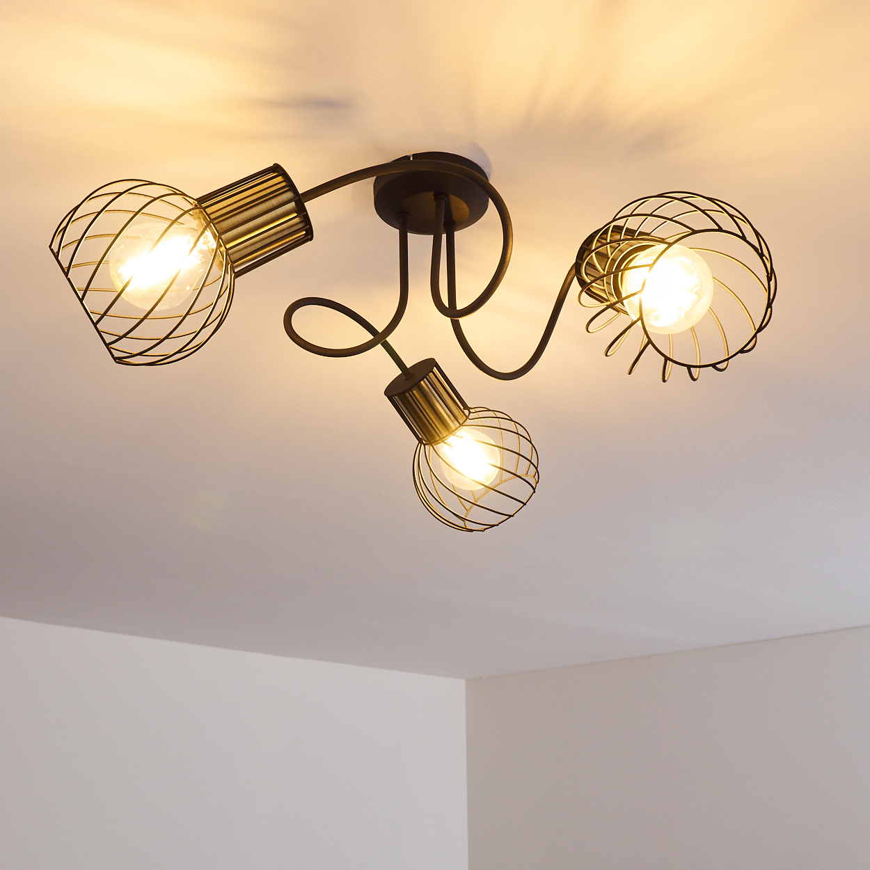 Eine Deckenleuchte mit drei Lampen. Die Lampen sind in schwarzen Drahtrahmen eingeschlossen. Das Licht scheint in einen weißen Deckenraum.