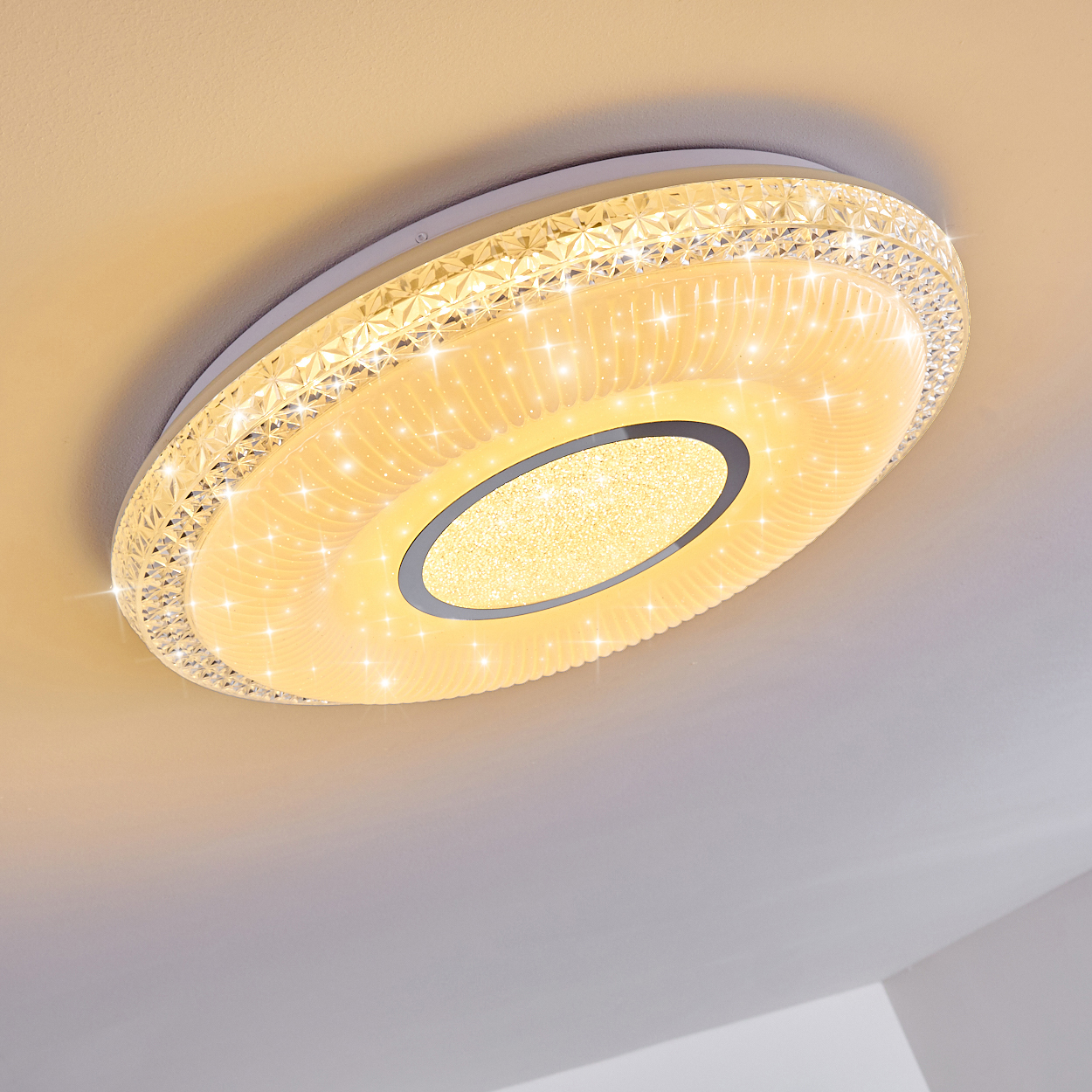 Eine moderne, beleuchtete Deckenleuchte. Sie hat einen kristallartigen Rand und ein zentrales Oval mit Glitzer, vor einer weißen Decke.