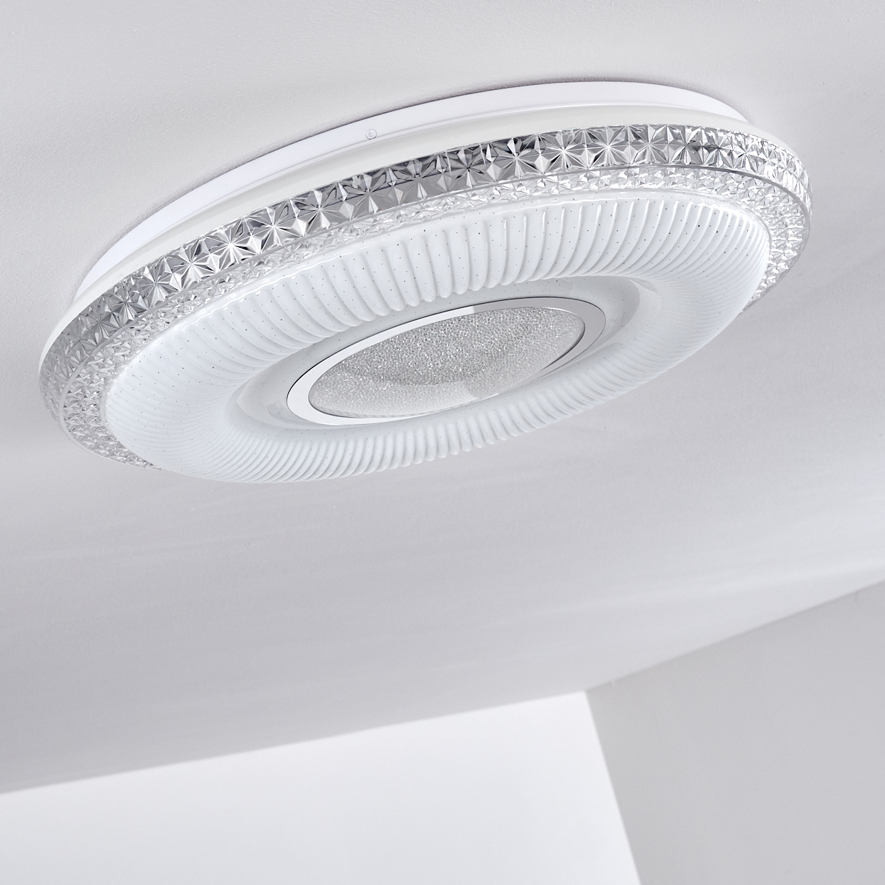 Eine weiße, kreisförmige Deckenleuchte mit strukturiertem Design und einem glitzernden, silbernen Zentrum, vor einer weißen Decke.