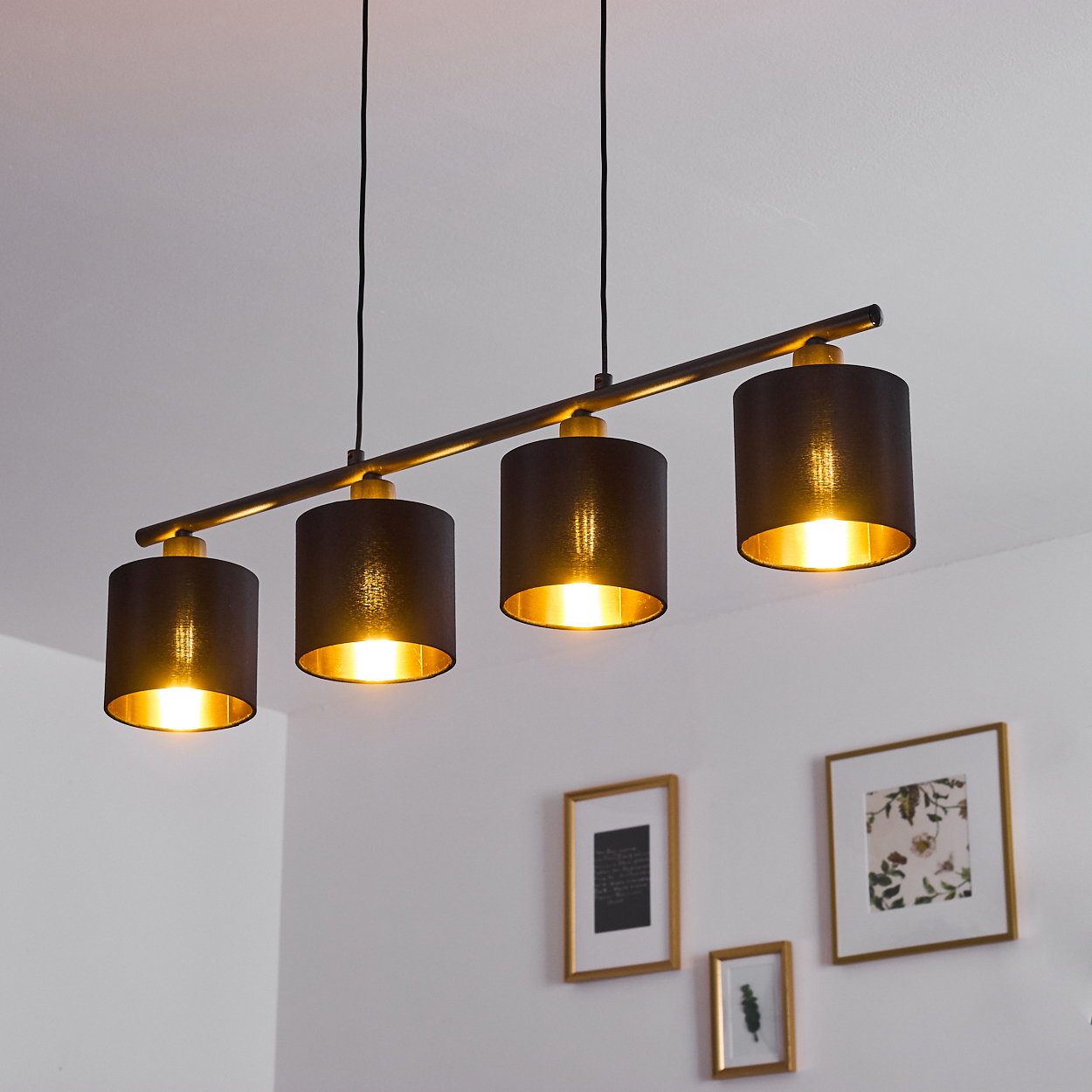 Eine moderne Deckenlampe mit vier schwarzen Schirmen, goldenen Innenräumen und hellem Licht. Weiße Wände mit gerahmten Bildern im Hintergrund.