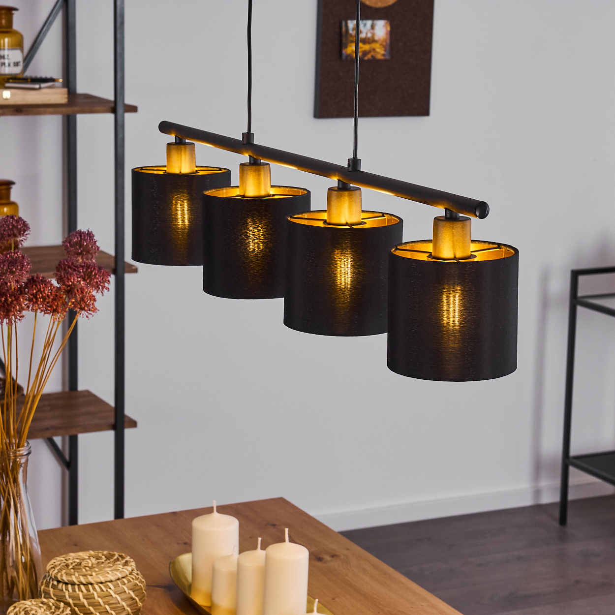 Eine schwarze, moderne Leuchte mit vier Lampenschirmen hängt über einem Holztisch mit Kerzen und einer Blumenvase.