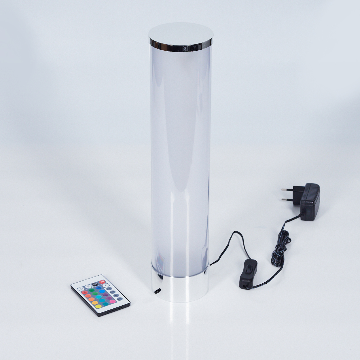 Eine zylindrische LED-Lampe mit Fernbedienung, Netzteil und Schalter auf weißer Oberfläche. Die Lampe ist silber und klar.