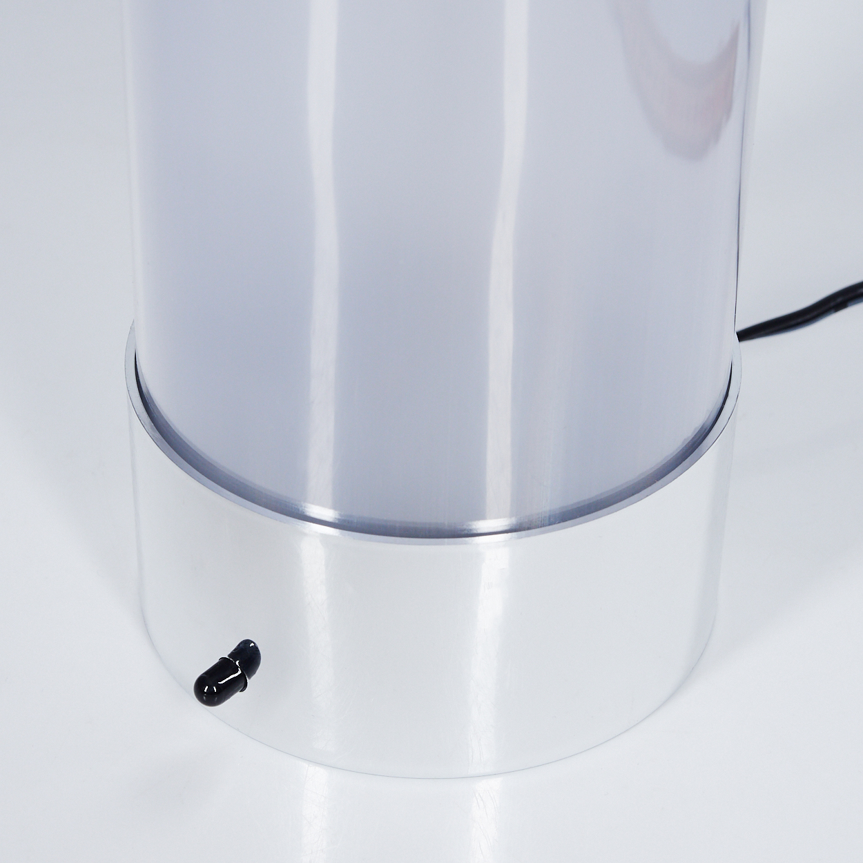 Eine zylindrische Lampe mit silbernem Sockel und einem durchscheinenden, lichtstreuenden oberen Bereich. Ein schwarzes Netzkabel geht von der Seite aus.