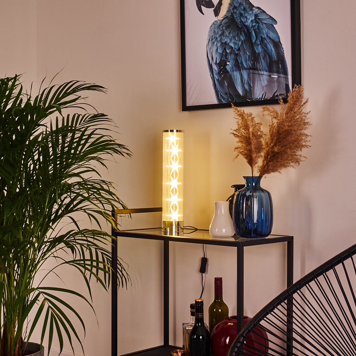 Eine beleuchtete sternförmige Lampe und eine blaue Vase mit trockenen Pflanzen stehen auf einem Tisch. Ein gerahmtes Papageienbild hängt an der Wand.
