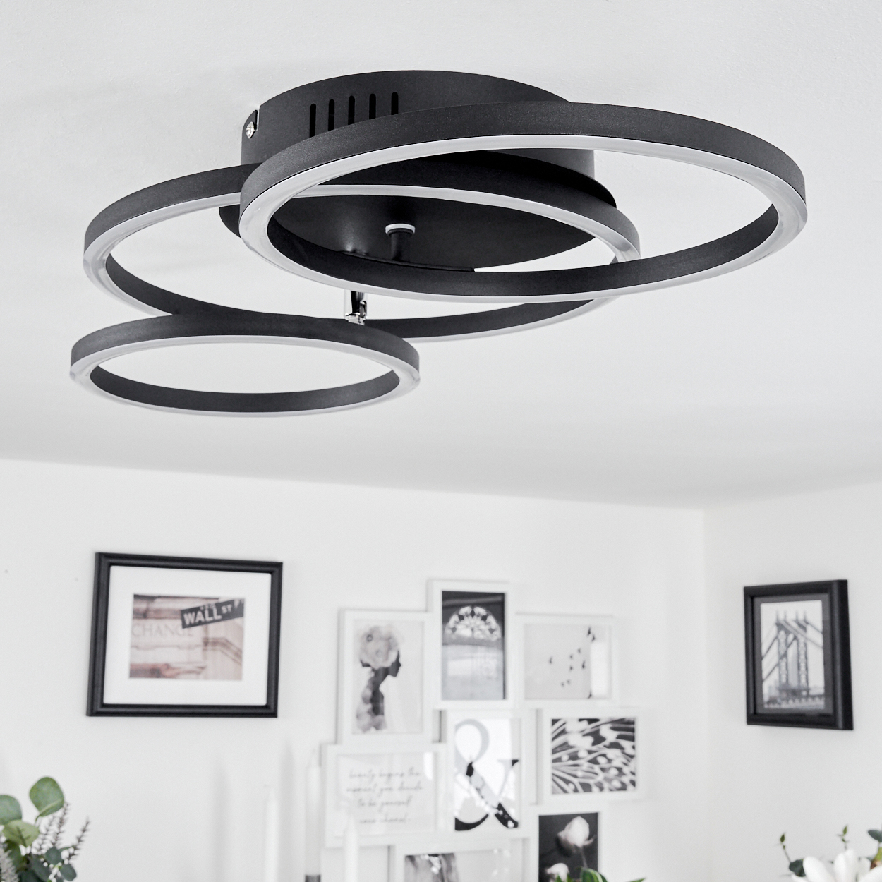 Eine moderne Deckenleuchte mit drei sich überlappenden schwarzen und silbernen Ringen. Die weiße Decke und Wand zeigen gerahmte Kunst.