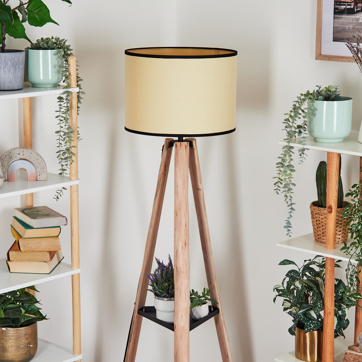 Eine Holz-Dreibein-Lampe mit cremefarbenem Lampenschirm. Auf Regalen stehen Pflanzen und Bücher. Der Hintergrund ist eine weiße Wand.