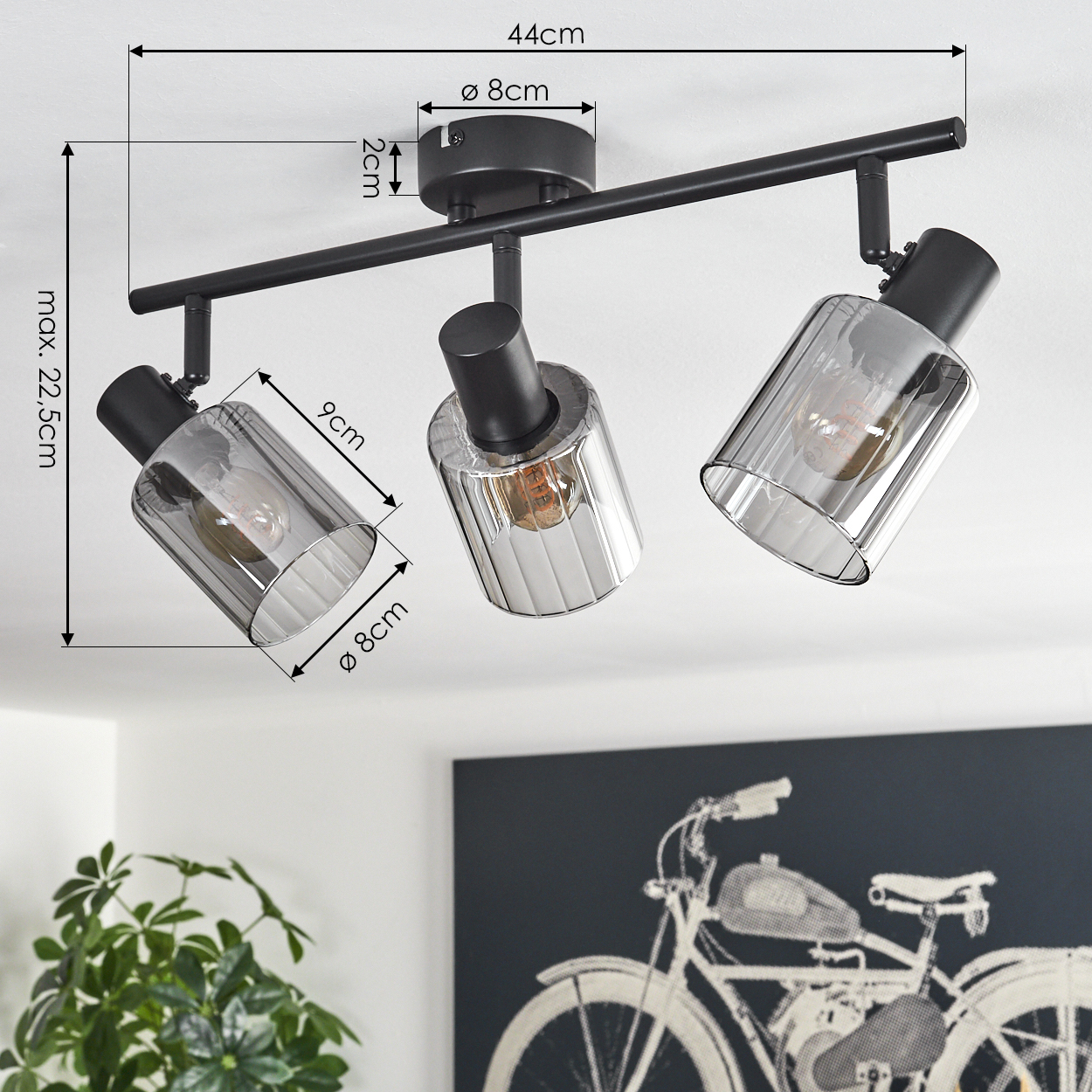 Eine schwarze Deckenleuchte mit drei verstellbaren Lampen. Maße sind beschriftet. Pflanze und Fahrrad-Kunstwerk sind im Hintergrund sichtbar.