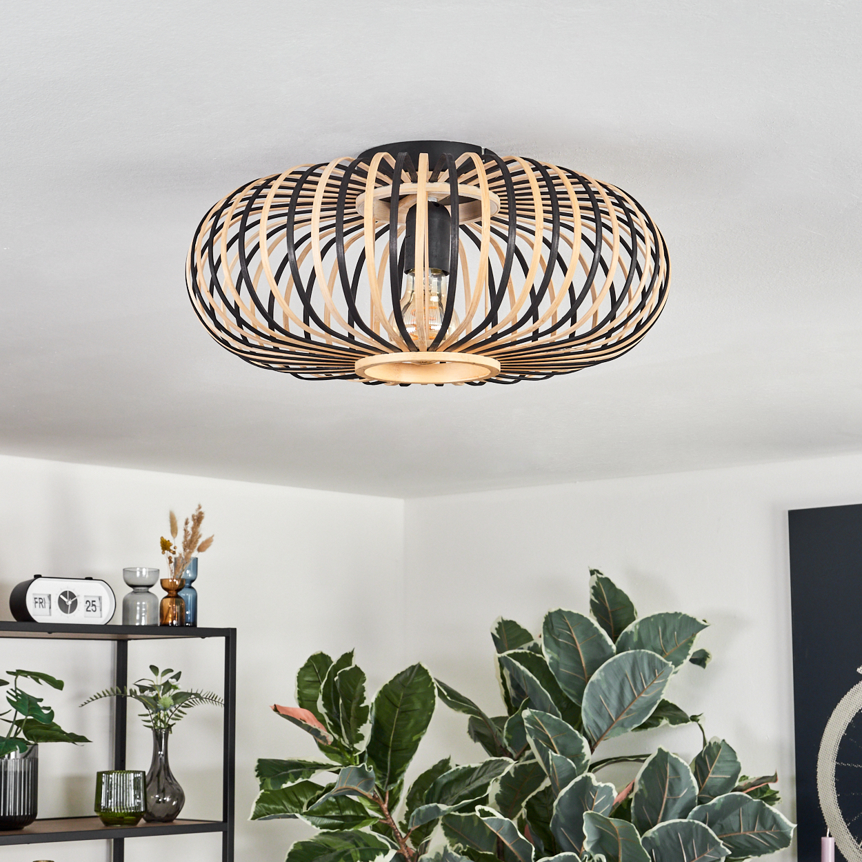 Eine hölzerne Deckenlampe mit schwarz-beigen Streifen. Die Lampe befindet sich in einem weißen Raum, mit Pflanzen und einem Regal im Vordergrund.