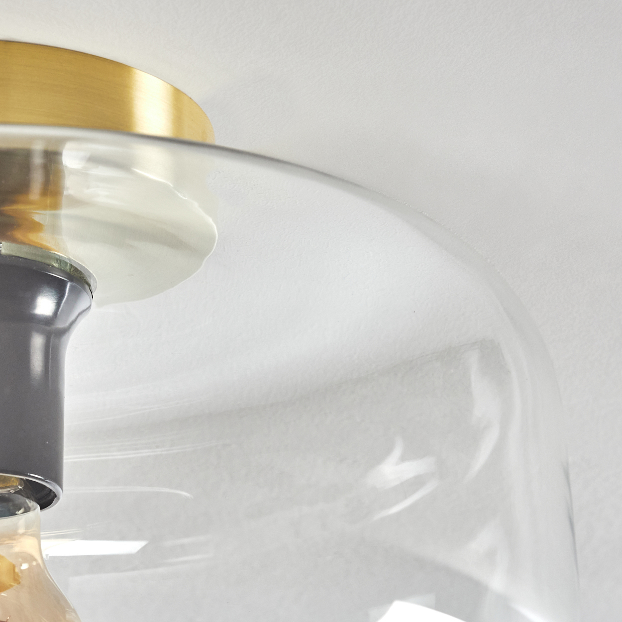 Nahaufnahme einer modernen Leuchte. Sie hat einen goldfarbenen Aufsatz und einen transparenten Glasschirm. Der Hintergrund ist eine weiße Wand.