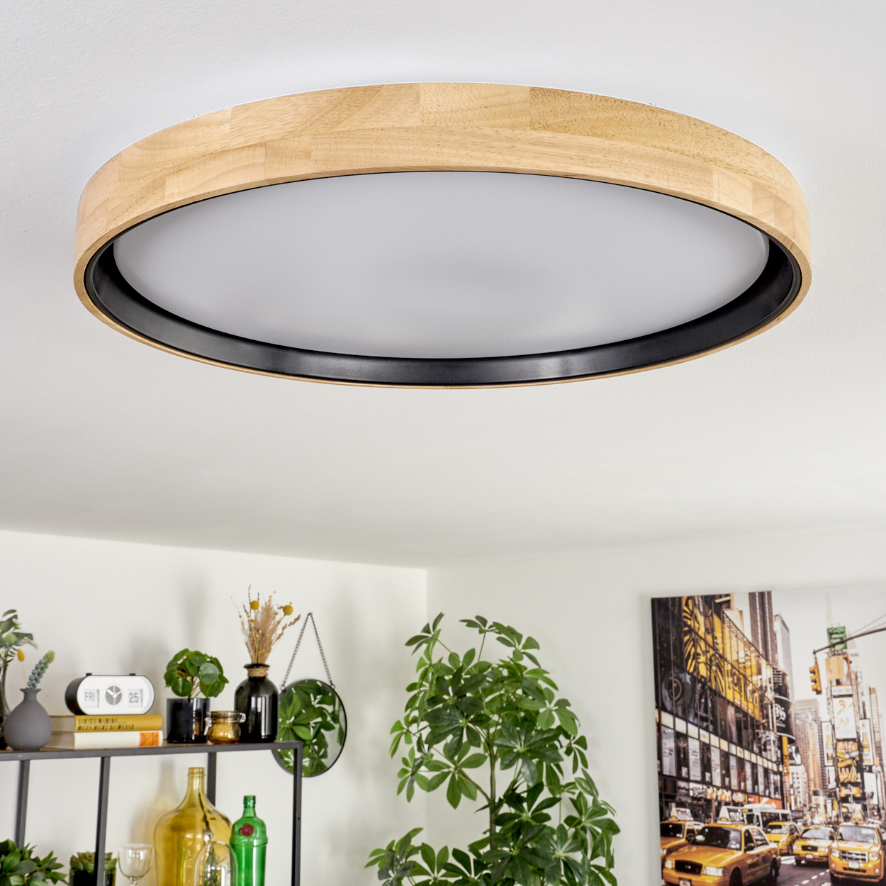 Eine kreisförmige Deckenlampe aus Holz. Darunter ein Regal mit Dekoration und Pflanzen. Rechts hängt ein Stadtbild.