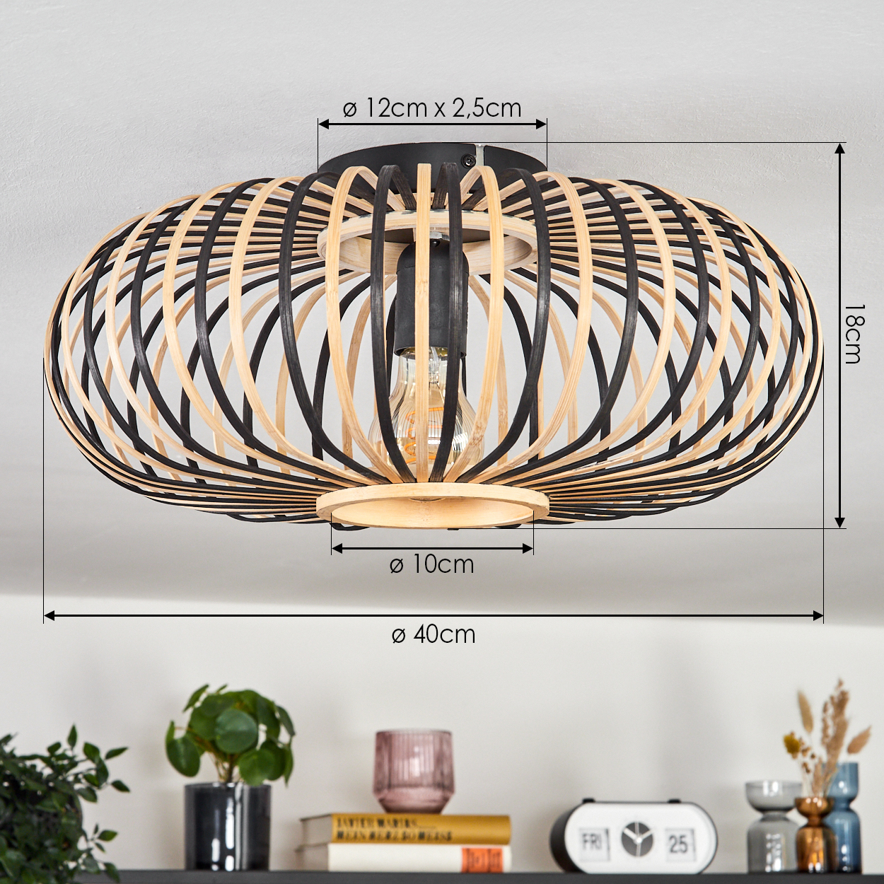 Eine Deckenlampe mit einem schwarz-beigen Holzrahmen. Abmessungen sind angegeben. Ein Regal darunter enthält Pflanzen, Bücher und Dekorationsartikel.