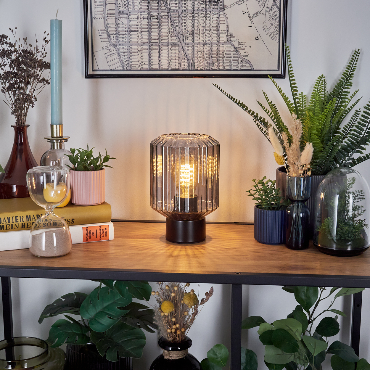 Eine Lampe mit einer brennenden Glühbirne steht auf einem Holzregal mit Büchern und Pflanzen. Darüber hängt eine Karte. Weitere Pflanzen und Dekorationsartikel sind vorhanden.