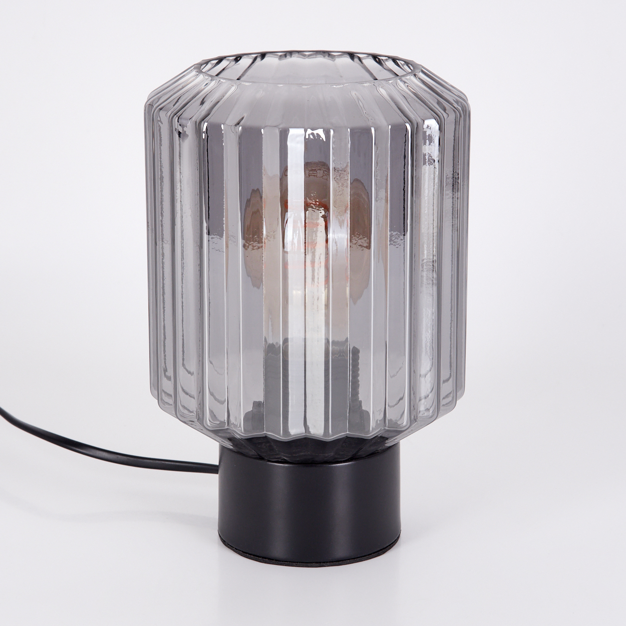 Eine graue Glaslampe mit schwarzem Sockel und sichtbarer Glühbirne. Die Lampe steht auf einer weißen Oberfläche.