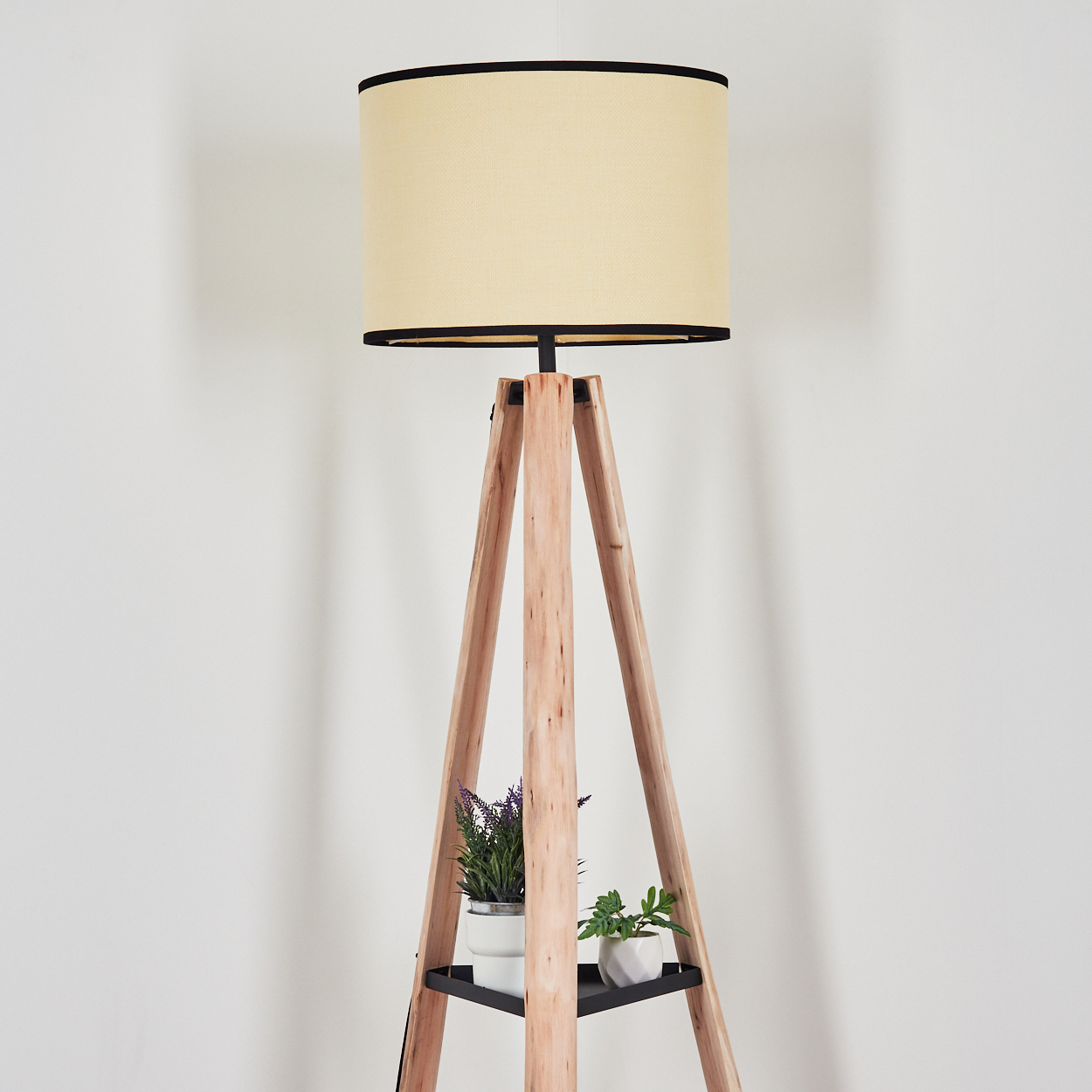 Eine hölzerne Stativ-Stehlampe mit beigefarbenem Lampenschirm. Zwei kleine Topfpflanzen stehen auf einem Regal. Weiße Wand im Hintergrund.