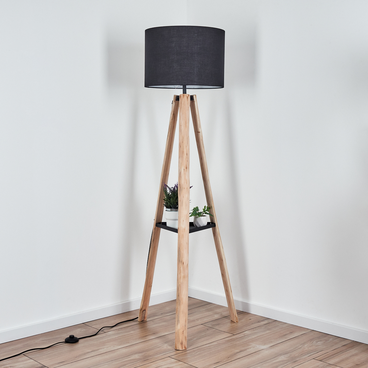 Eine hölzerne Stativ-Stehlampe mit schwarzem Lampenschirm. Sie hat ein Regal mit Pflanzen. Sie steht vor einer weißen Wand, mit Kabel und Schalter.