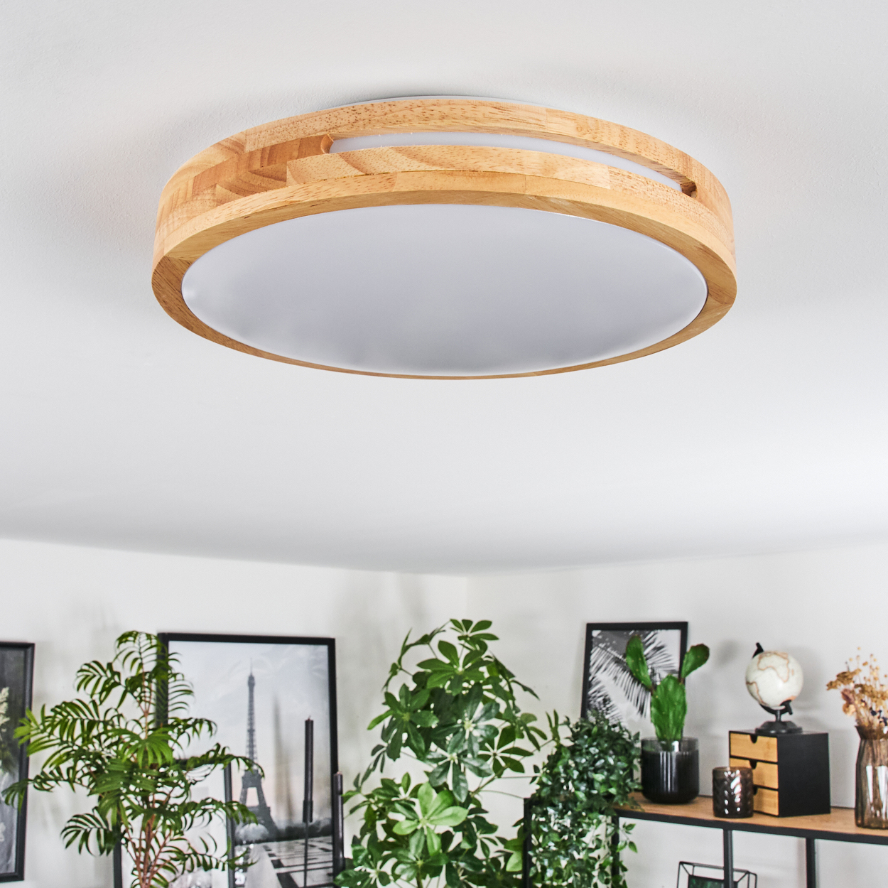 Eine Deckenleuchte aus Holz. Darunter schmücken Pflanzen und gerahmte Bilder einen Raum. Das Licht ist kreisförmig mit weißem Schirm.