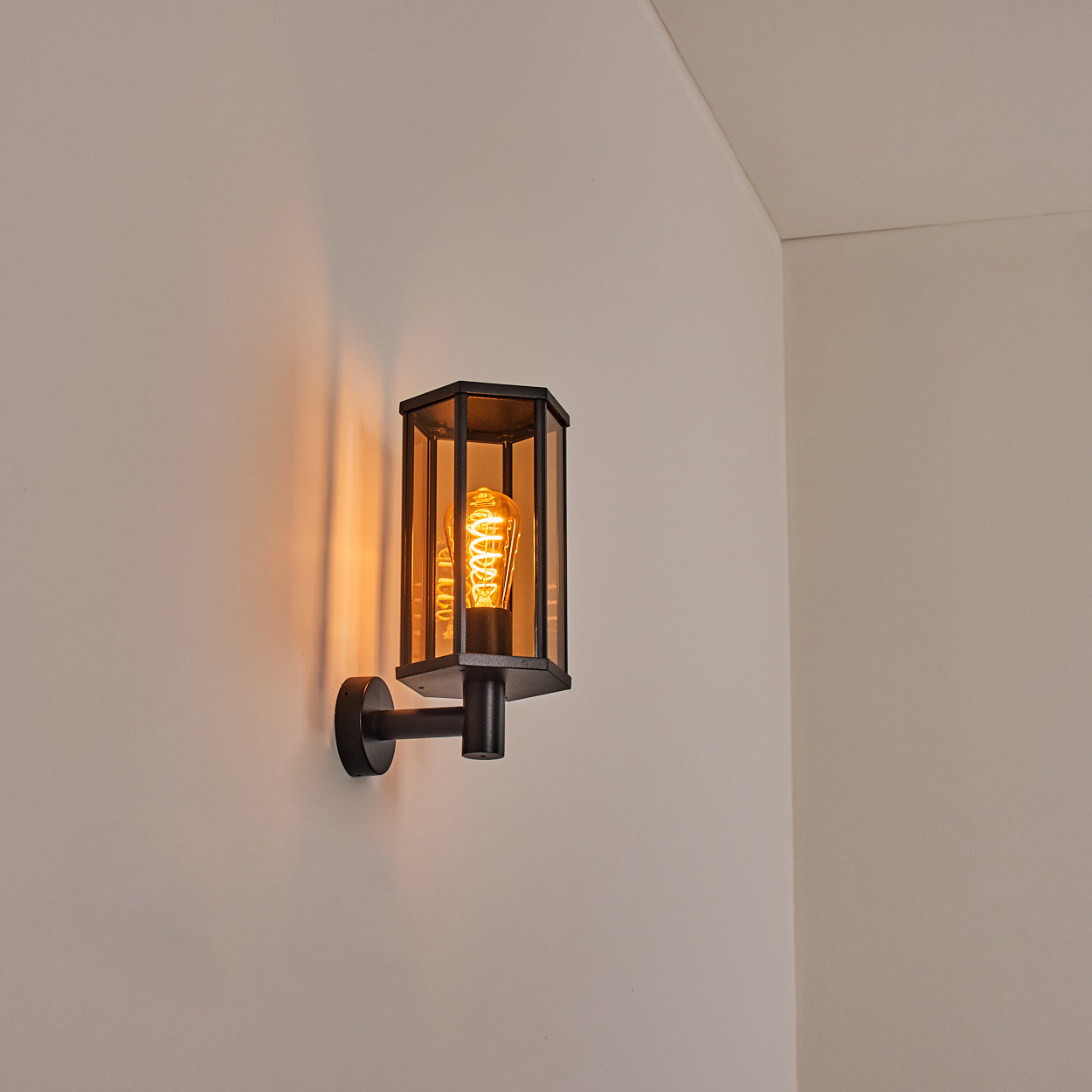 Eine schwarze Wandlampe mit leuchtender Glühbirne ist an einer weißen Wand befestigt. Die Lampe hat ein geometrisches Design mit Glaspaneelen.