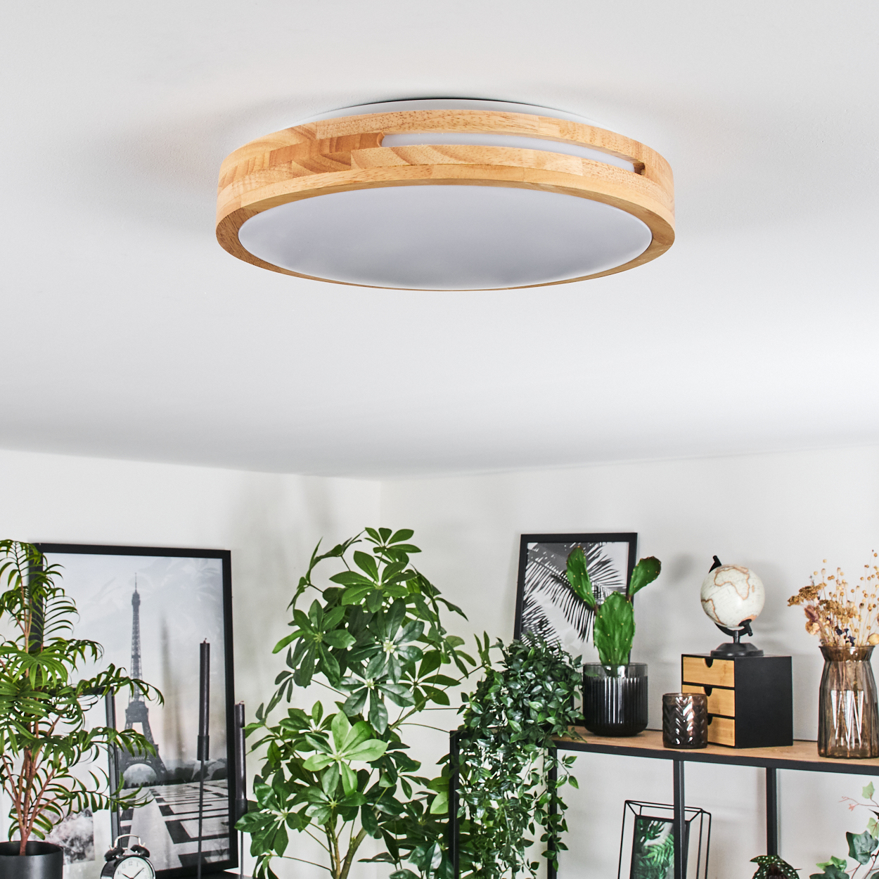 Eine Deckenlampe aus Holz ist zentriert. Darunter befinden sich grüne Pflanzen, Bilder und Dekorationsartikel auf einem Regal. Weiße Wände und Decke.
