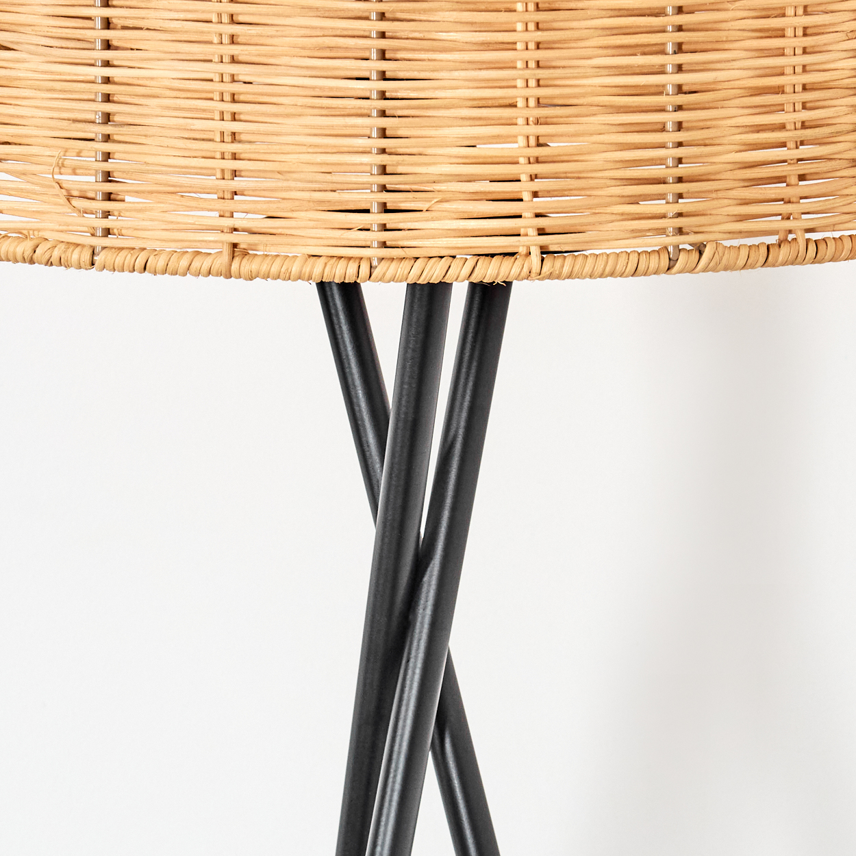 Nahaufnahme einer Lampe. Geflochtener Rattanschatten, schwarze Metallbeine kreuzen sich. Weißer Hintergrund. Hellbraune und schwarze Farben.