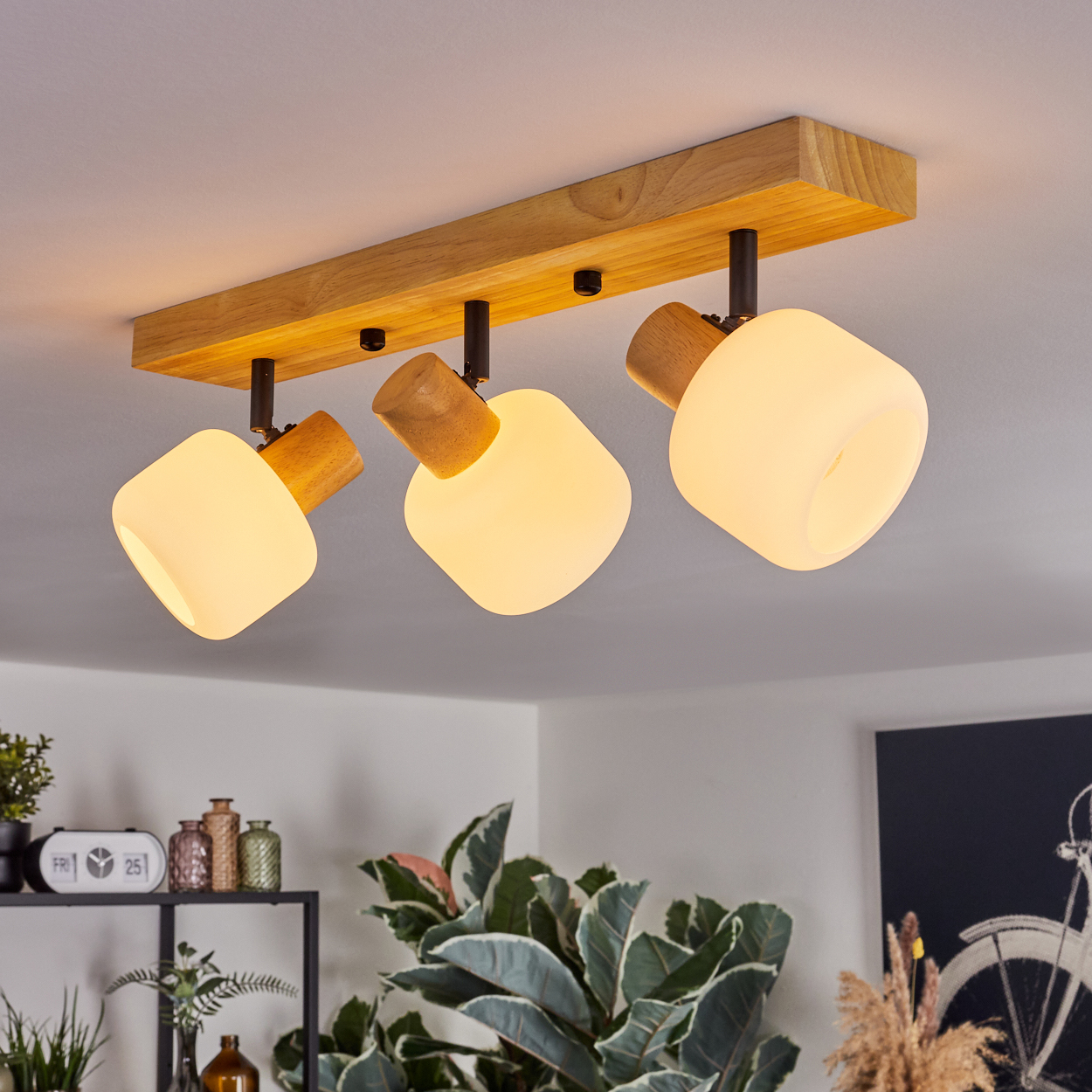 Eine Deckenleuchte aus Holz mit drei Lampen. Jede Lampe hat einen Holzsockel und eine weiße, abgerundete Leuchte. Pflanzen und Dekoration im Hintergrund.