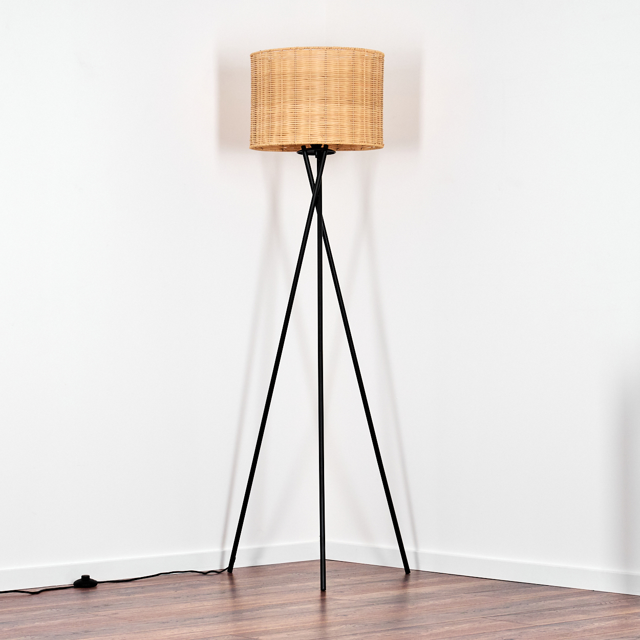 Eine Stehlampe mit gewebtem Lampenschirm steht in einer Ecke. Sie hat ein schwarzes Stativ. Der Boden ist Holz, die Wände sind weiß.